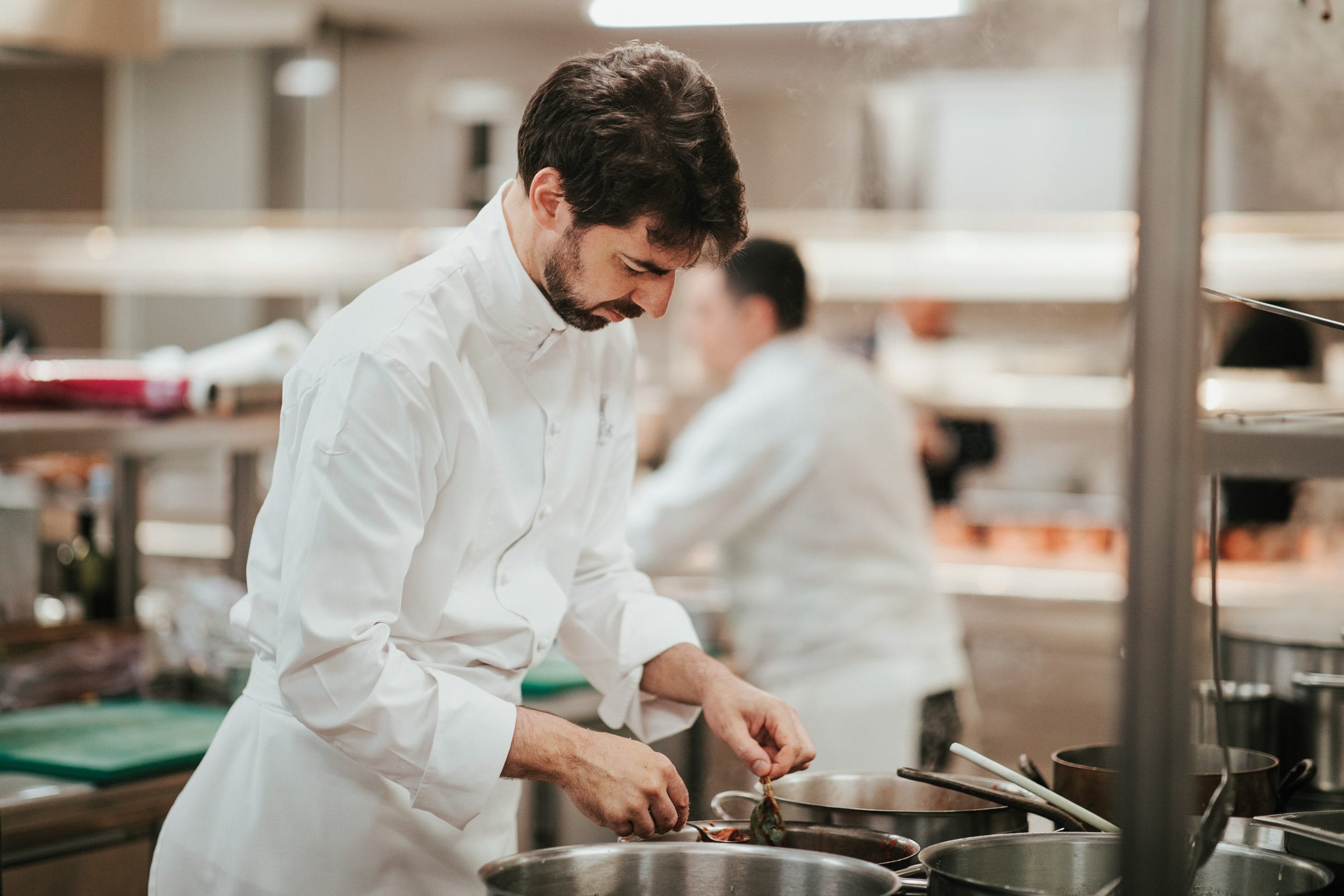 Chef preparing a dish in a professional kitchen, focusing intently on his work with various cooking utensils around him.