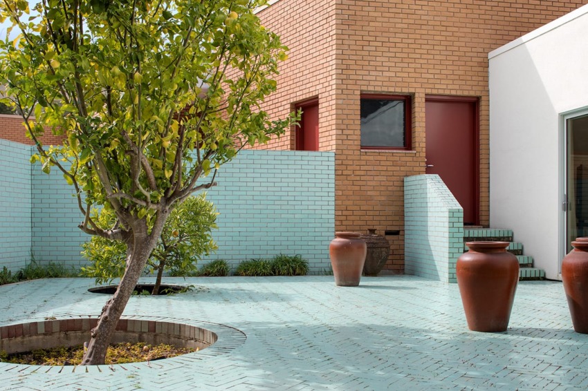 Modern courtyard with light blue brick floor, terracotta pots, and a lemon tree, adjacent to a brick and white building.