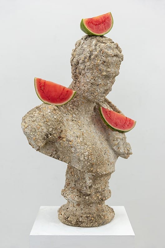 Stone bust adorned with watermelon slices on head and shoulders, displayed on a white pedestal.