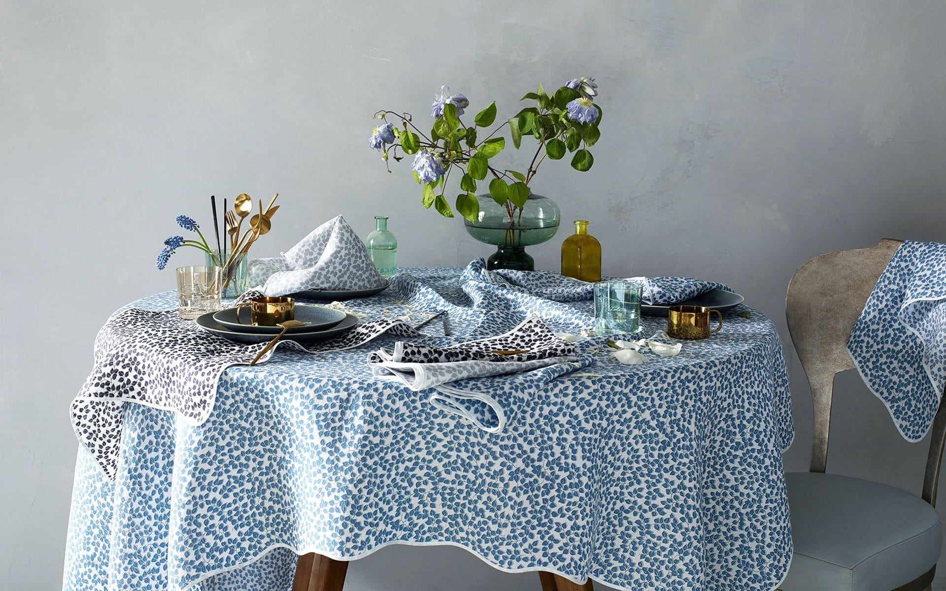 Elegant table setting with a blue patterned tablecloth, plates, cutlery, and a vase of flowers against a neutral background.