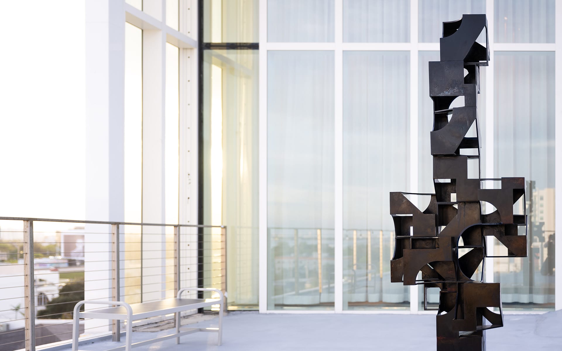 Outdoor abstract black metal sculpture on a modern balcony with glass walls and a city view in the background.