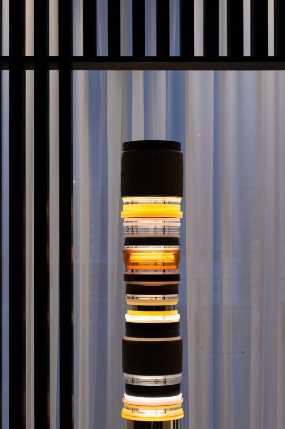 Modern floor lamp with stacked circular shades emitting warm light, set against sheer curtains and vertical slatted wall background.