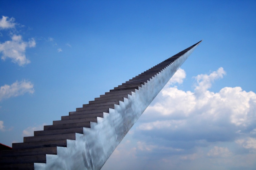 A staircase sculpture ascending into the blue sky with clouds in the background.