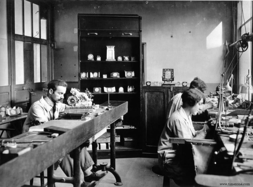 Watchmakers working at benches in a vintage workshop, surrounded by tools and clocks.