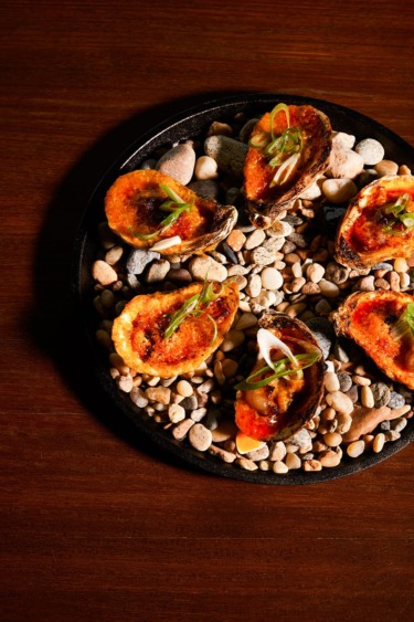 Grilled oysters on a bed of pebbles, garnished with green onions, served on a black plate on a wooden table.
