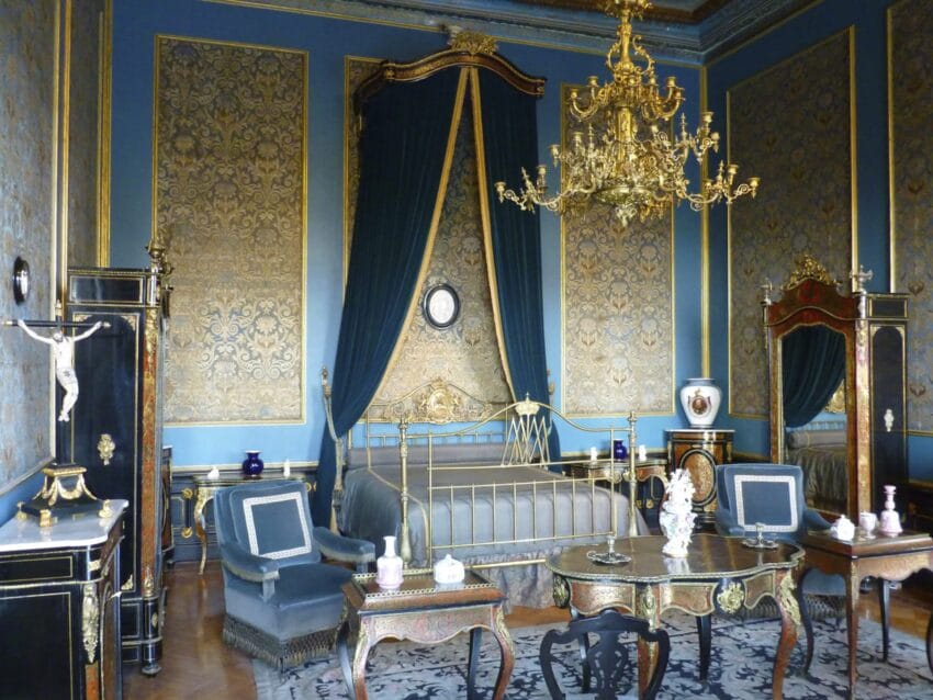 Luxurious antique bedroom with ornate gold details, blue walls, a decorative bed, and an elegant chandelier.