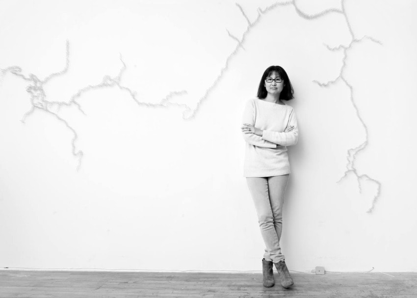 Person standing against a white wall with abstract line art, wearing a sweater and jeans, indoor setting, black and white photo