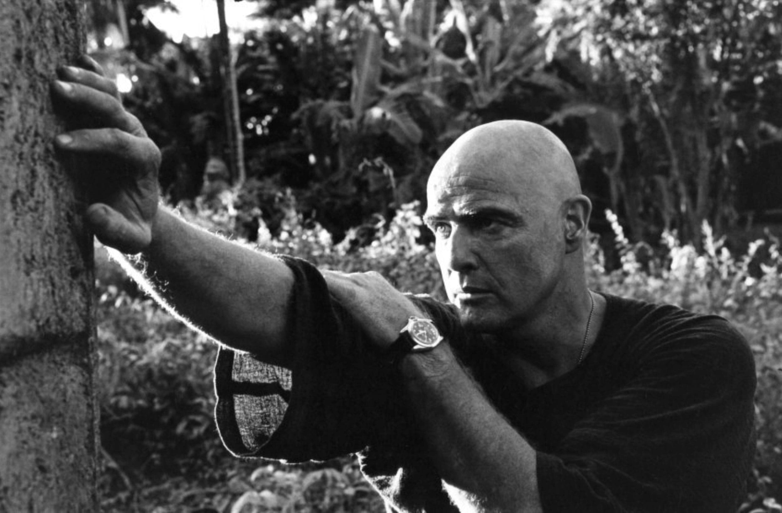Bald man in a dark shirt leaning against a tree outdoors, surrounded by dense tropical foliage.