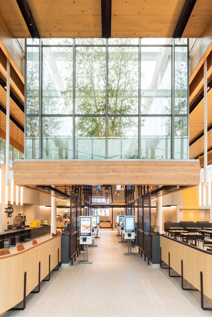 Modern restaurant interior with wooden accents, digital kiosks, tall glass windows, and a tree visible through the glass.
