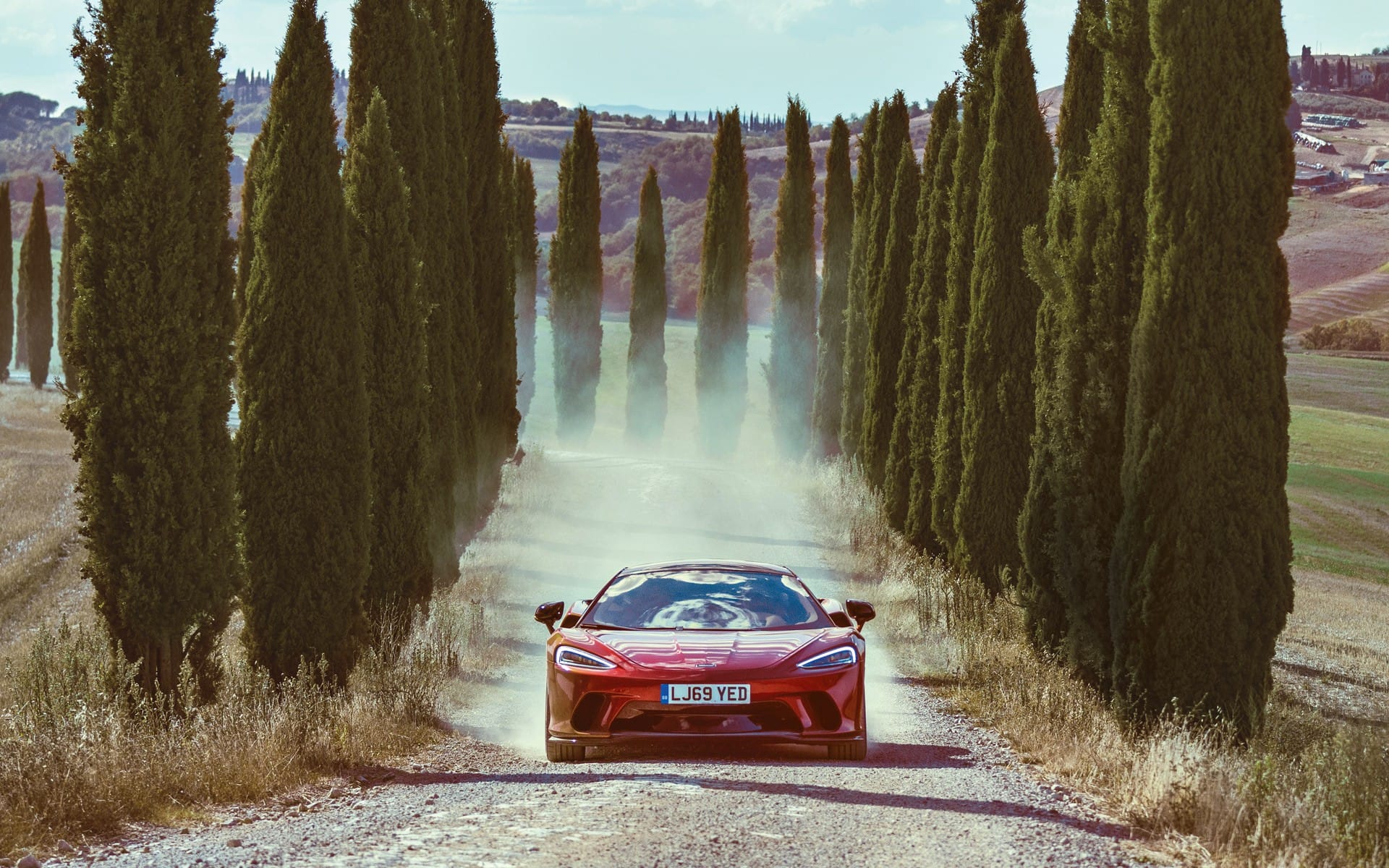 Red sports car driving on a dirt road lined with tall cypress trees under a clear blue sky.