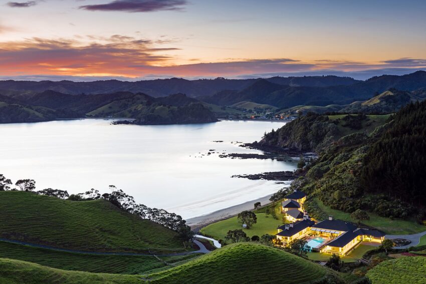 Scenic view of a coastal landscape with a luxury hillside villa, lush greenery, and a vibrant sunset over calm waters.