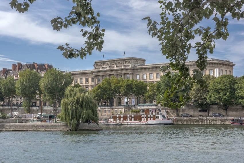 Riverboat cruising on the Seine with trees and buildings in the background, under a partly cloudy sky.