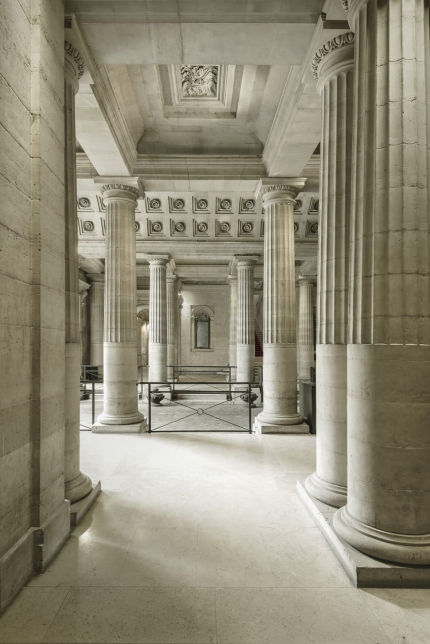 Interior of a grand building with large stone columns, decorated ceiling, and soft natural lighting.