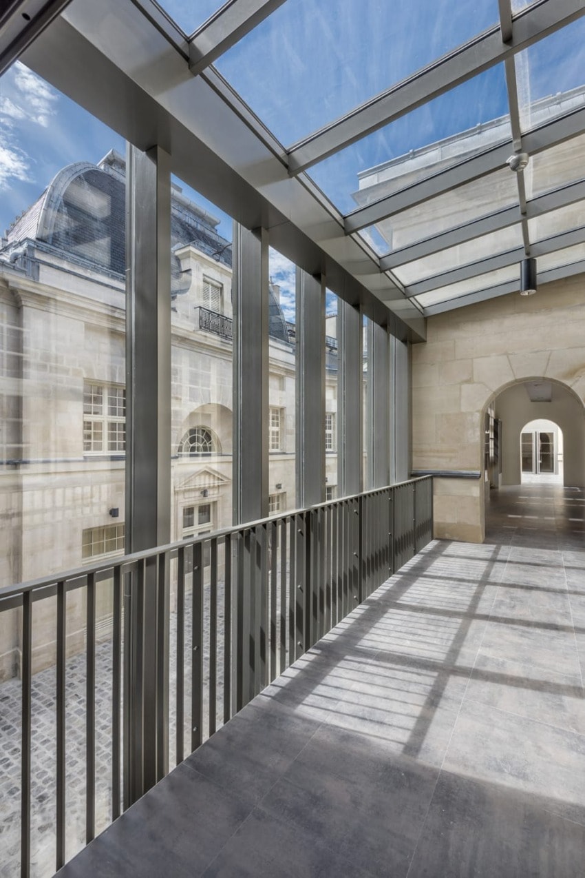 Modern glass walkway with metal railings overlooking stone building and courtyard on a sunny day.