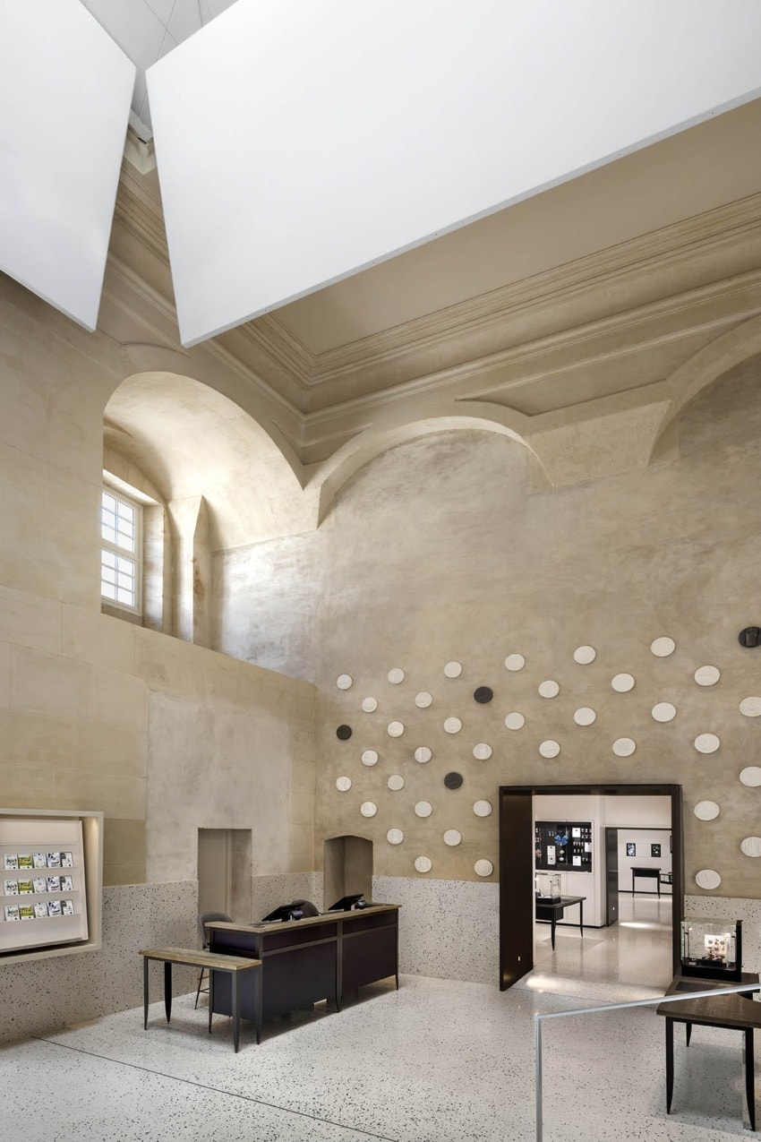 Modern museum interior with high ceiling, two black desks, dot-patterned wall, and a doorway leading to a brightly lit room.