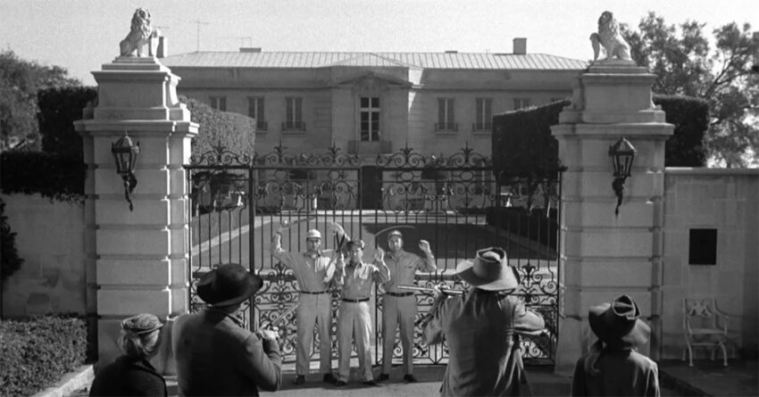 Group of soldiers guarding an ornate gate in front of a large mansion, with trees and statues on pillars nearby.