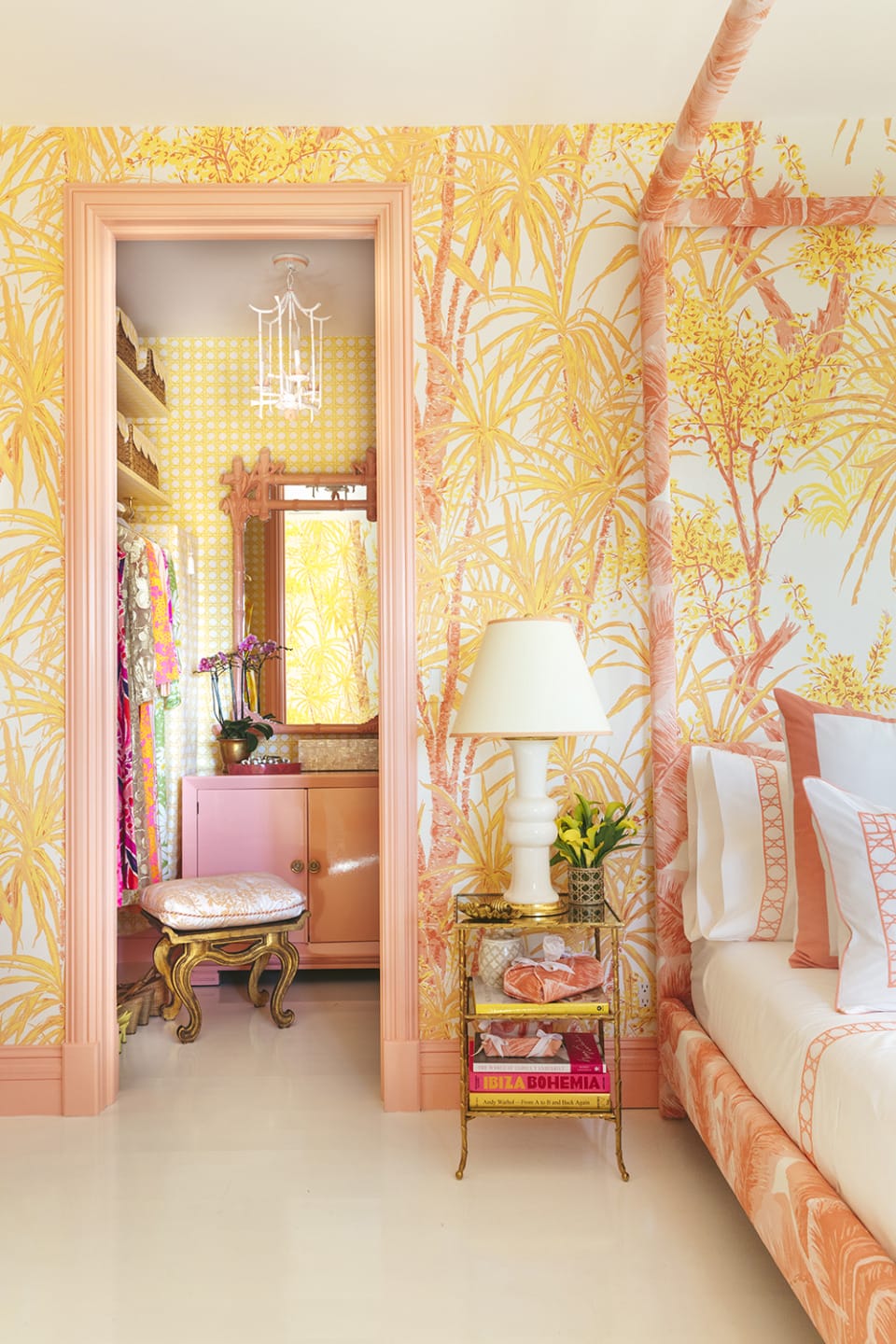 Vibrant bedroom with tropical-themed wallpaper, open closet, ornate stool, pink lamp, and stacked books on a bedside table.