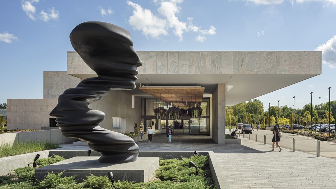 Modern art museum entrance with large abstract black sculpture, landscaped pathway, and visitors approaching under a partly cloudy sky.