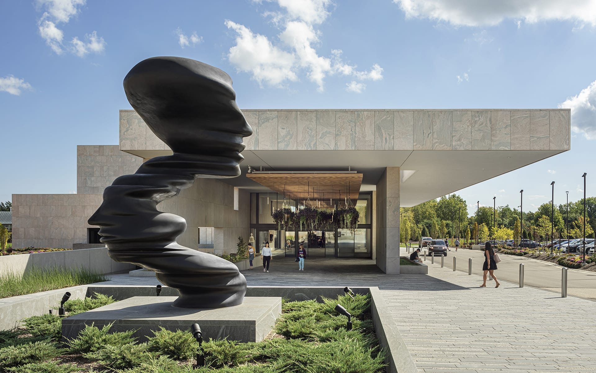 Modern art museum entrance with large abstract black sculpture, landscaped pathway, and visitors approaching under a partly cloudy sky.