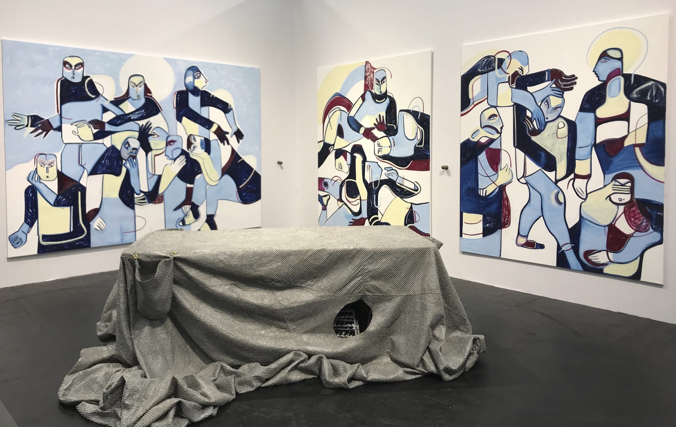 Abstract art exhibit featuring colorful geometric figures on three large canvases surrounding a draped table centerpiece.