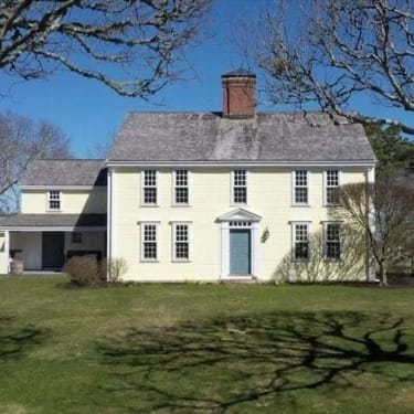 Historic two-story yellow house with a grey roof, surrounded by trees and a spacious green lawn under a clear blue sky.