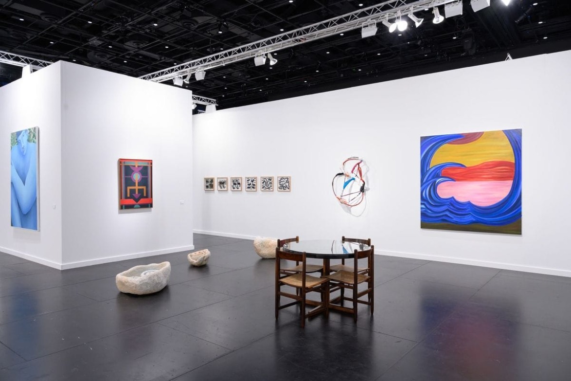 Art gallery exhibition featuring abstract paintings, sculptures, and a table with chairs in a modern, well-lit space