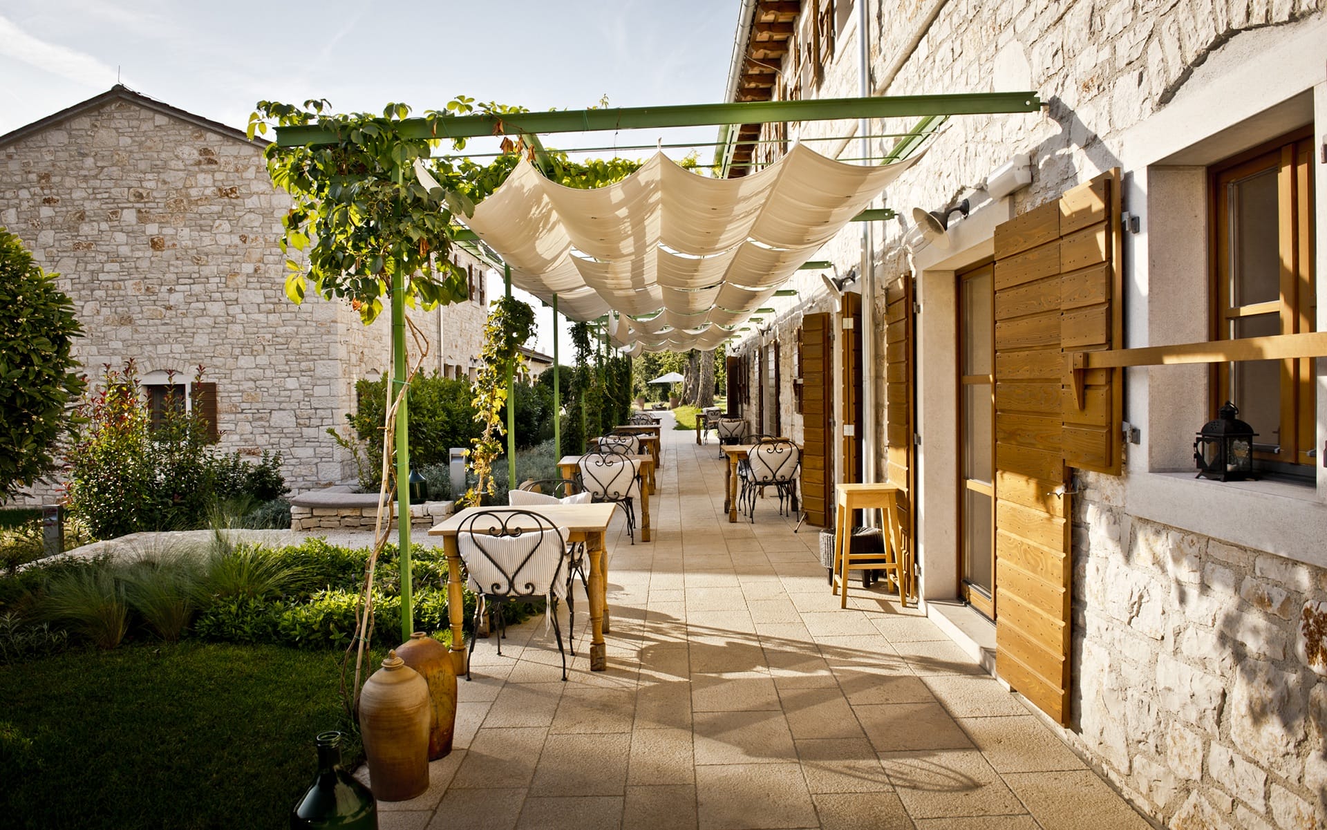 Rustic outdoor patio with stone walls, wooden tables, chairs, and a pergola canopy on a sunny day.