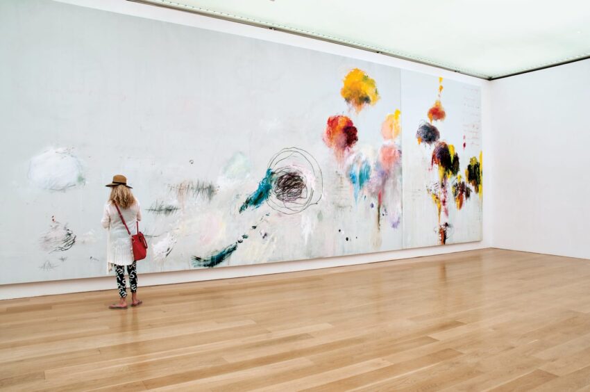 Person admiring a large abstract painting in a modern art gallery, featuring colorful strokes and dynamic patterns on a white wall.