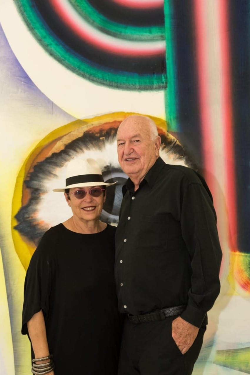 Two people smiling in front of a colorful abstract art background, one wearing a hat and sunglasses, both dressed in black.