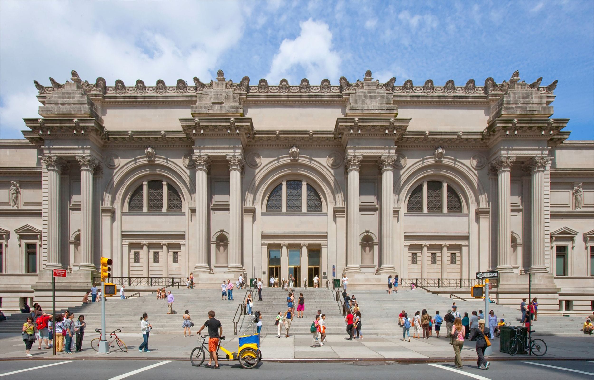 Exterior view of a large museum with a grand entrance and ornate architectural details, people walking in front.