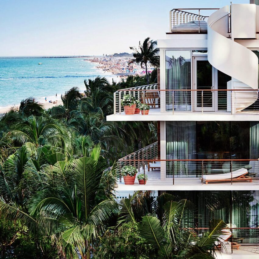 Beachfront modern building with glass balconies, spiral slide, surrounded by lush palm trees, overlooking a crowded beach.