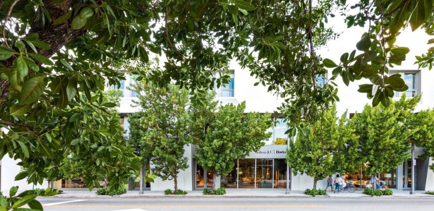 Street view of a white building framed by green trees and a sidewalk, with three people walking by the storefronts.