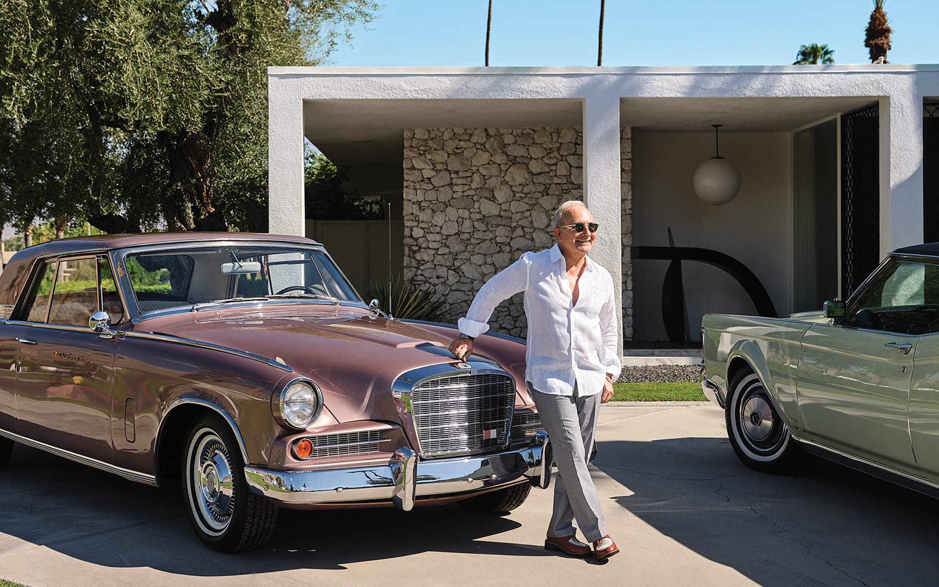 Man in sunglasses stands beside a vintage car in front of a modern house with a white exterior and stone accents.