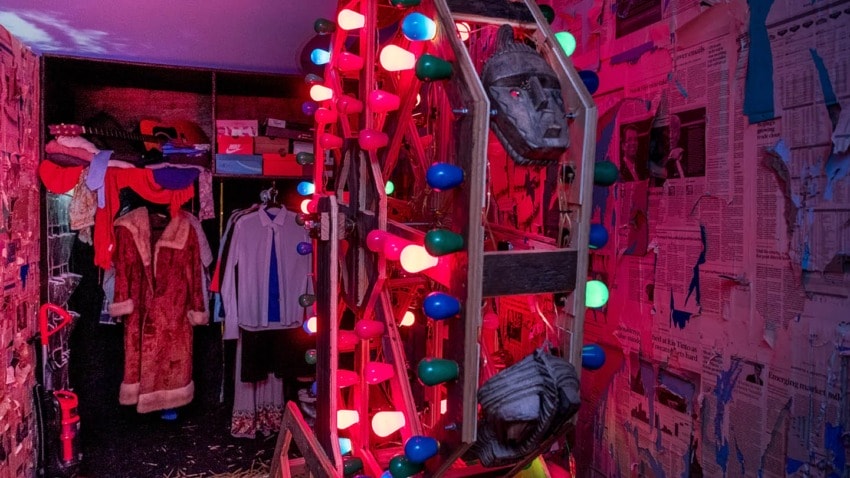 Colorful lights and masks on a wooden Ferris wheel in a decorated room with clothes and newspaper-covered walls.