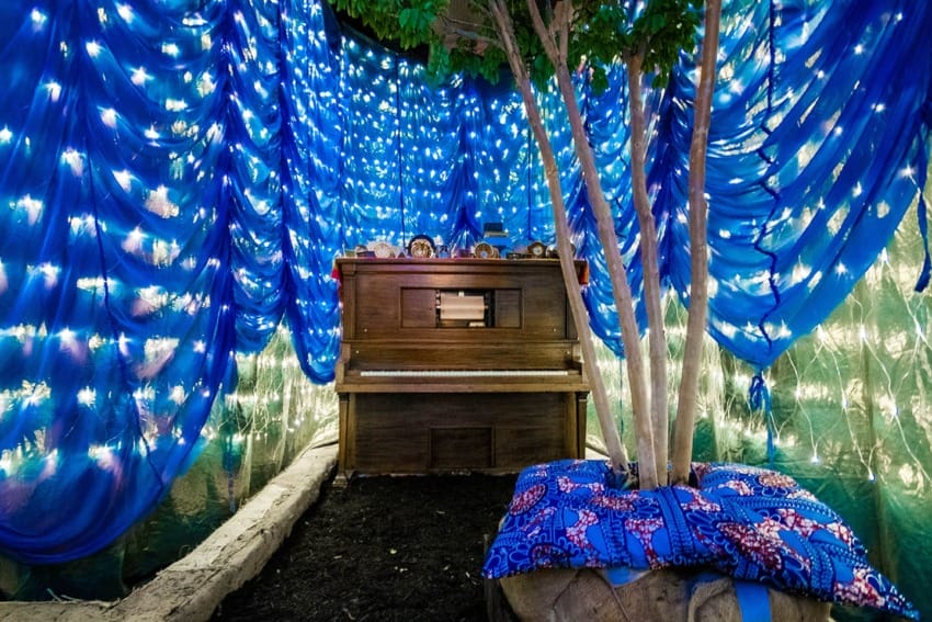 Piano under tree surrounded by blue curtains and string lights, creating a whimsical and magical atmosphere.