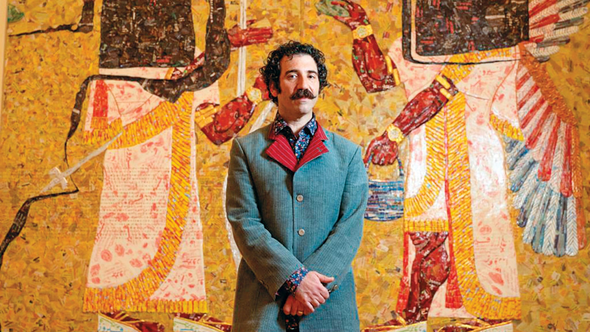 Man in a blue blazer with a red collar stands confidently in front of a vibrant, abstract yellow and red mosaic art piece.