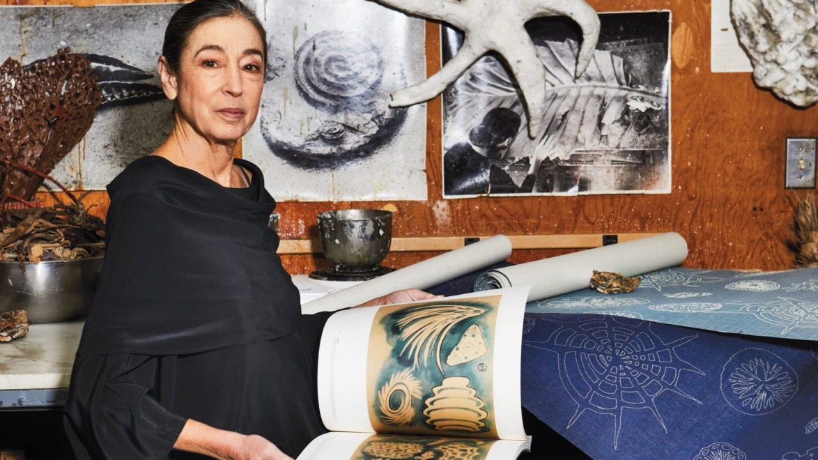 Artist with a book showcasing nature illustrations in a studio filled with art and scientific specimens.