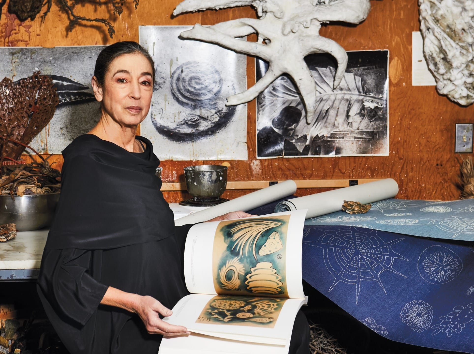 Artist with a book showcasing nature illustrations in a studio filled with art and scientific specimens.