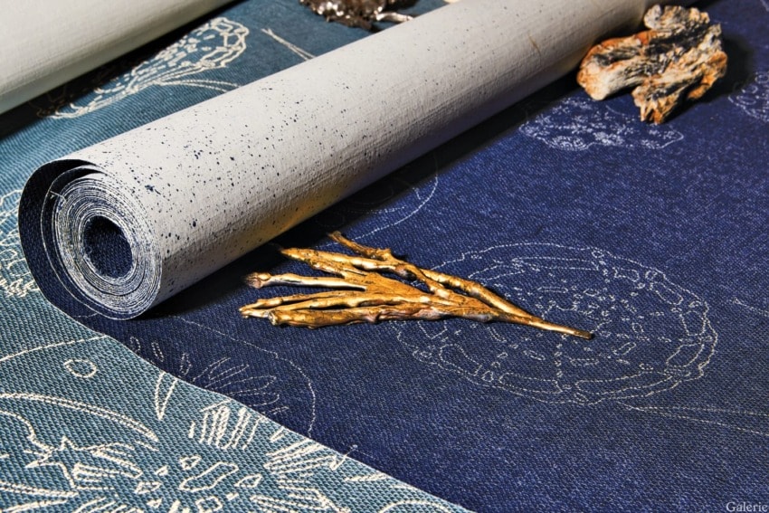 Rolled wallpaper on blue fabric with gold twigs and stones, showcasing a detailed artistic design and texture.