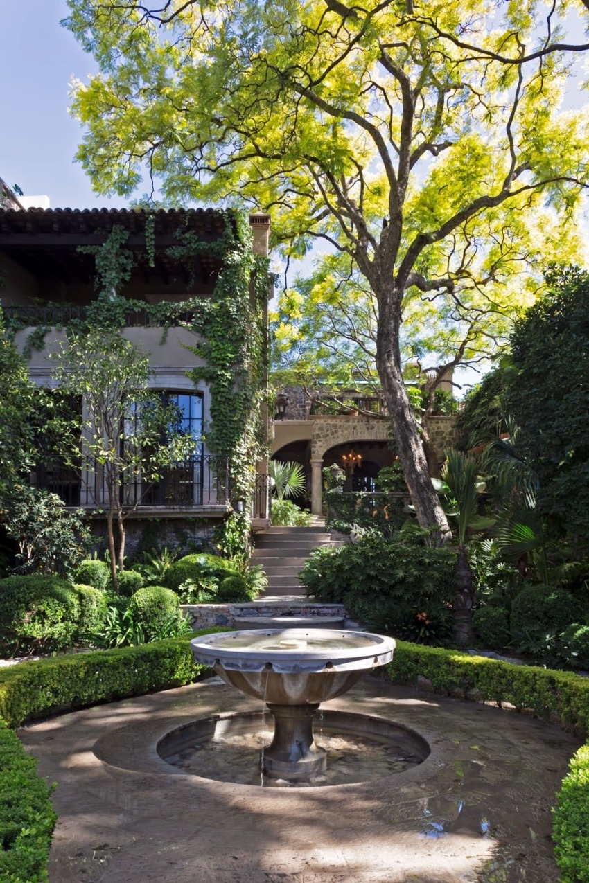 Courtyard garden with stone fountain, surrounded by lush greenery and a large tree under a bright sky.
