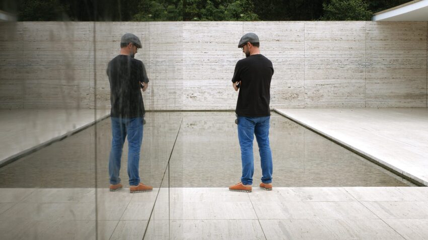 Man in jeans and cap standing in front of a reflective water feature with beige stone walls and greenery in the background.