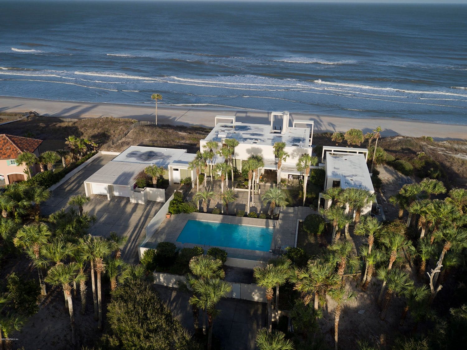 Aerial view of a beachfront house with palm trees, a large pool, and a wide sandy beach by the ocean.
