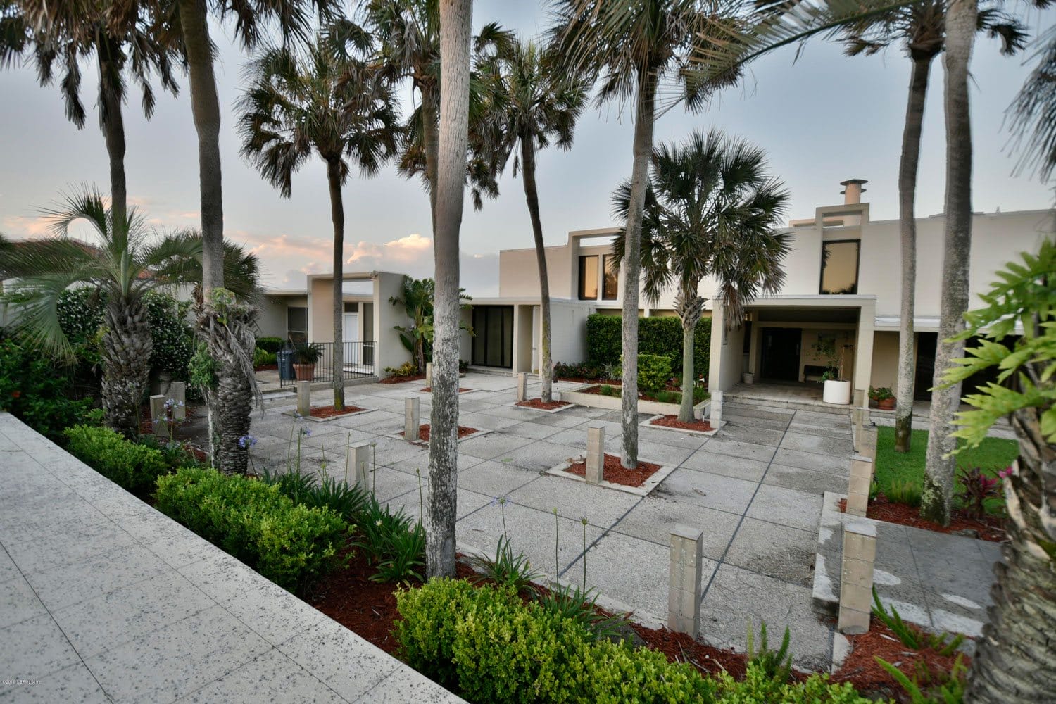 Modern coastal house with palm trees, large windows, and a shaded driveway surrounded by greenery at sunset.