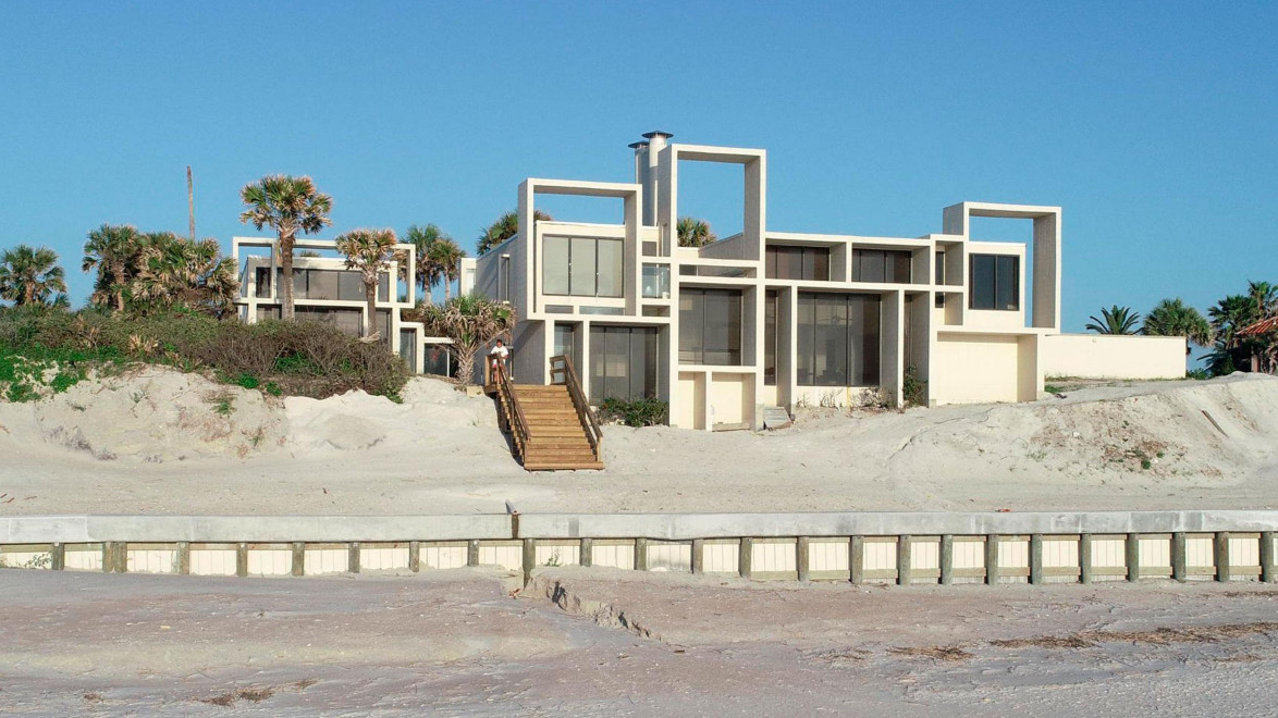 Modern beachfront house with geometric design, large windows, and palm trees on a sandy dune under a clear blue sky.