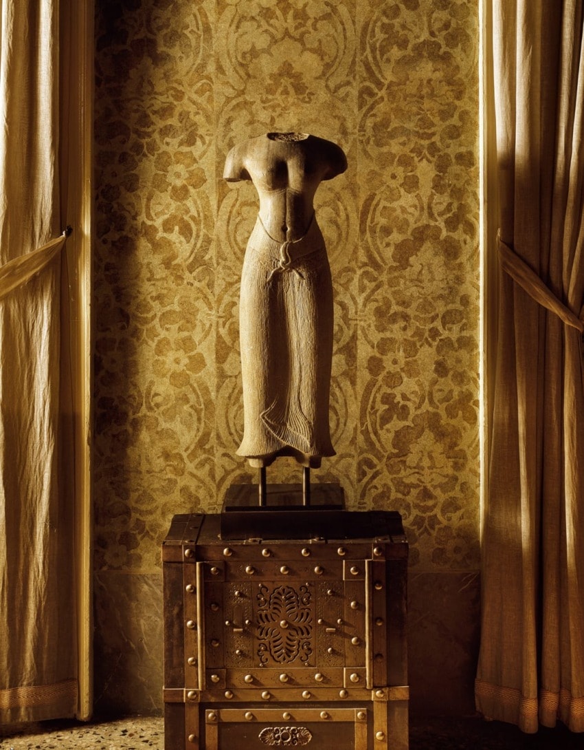 Ancient torso sculpture displayed on ornate chest, surrounded by luxurious drapery and patterned wallpaper in warm lighting.