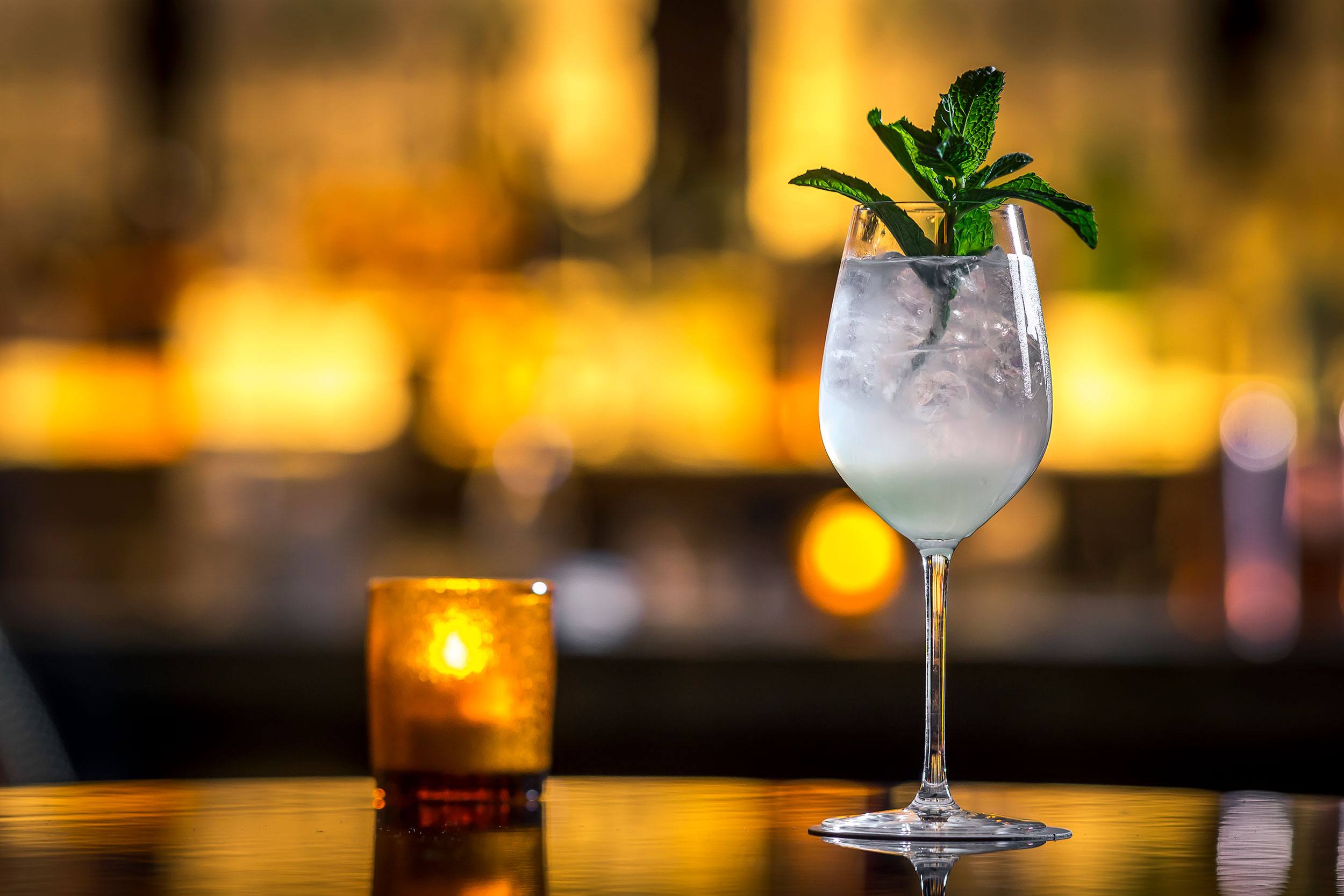 A cocktail glass with a mint garnish on a bar counter next to a lit candle in a dimly lit setting.