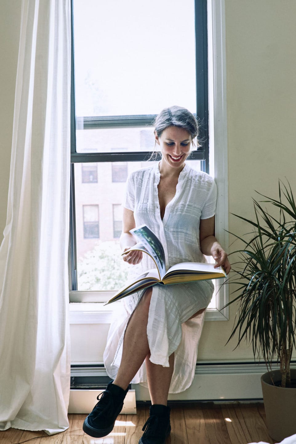 Woman in white dress reading a book while sitting by a window with a plant nearby.