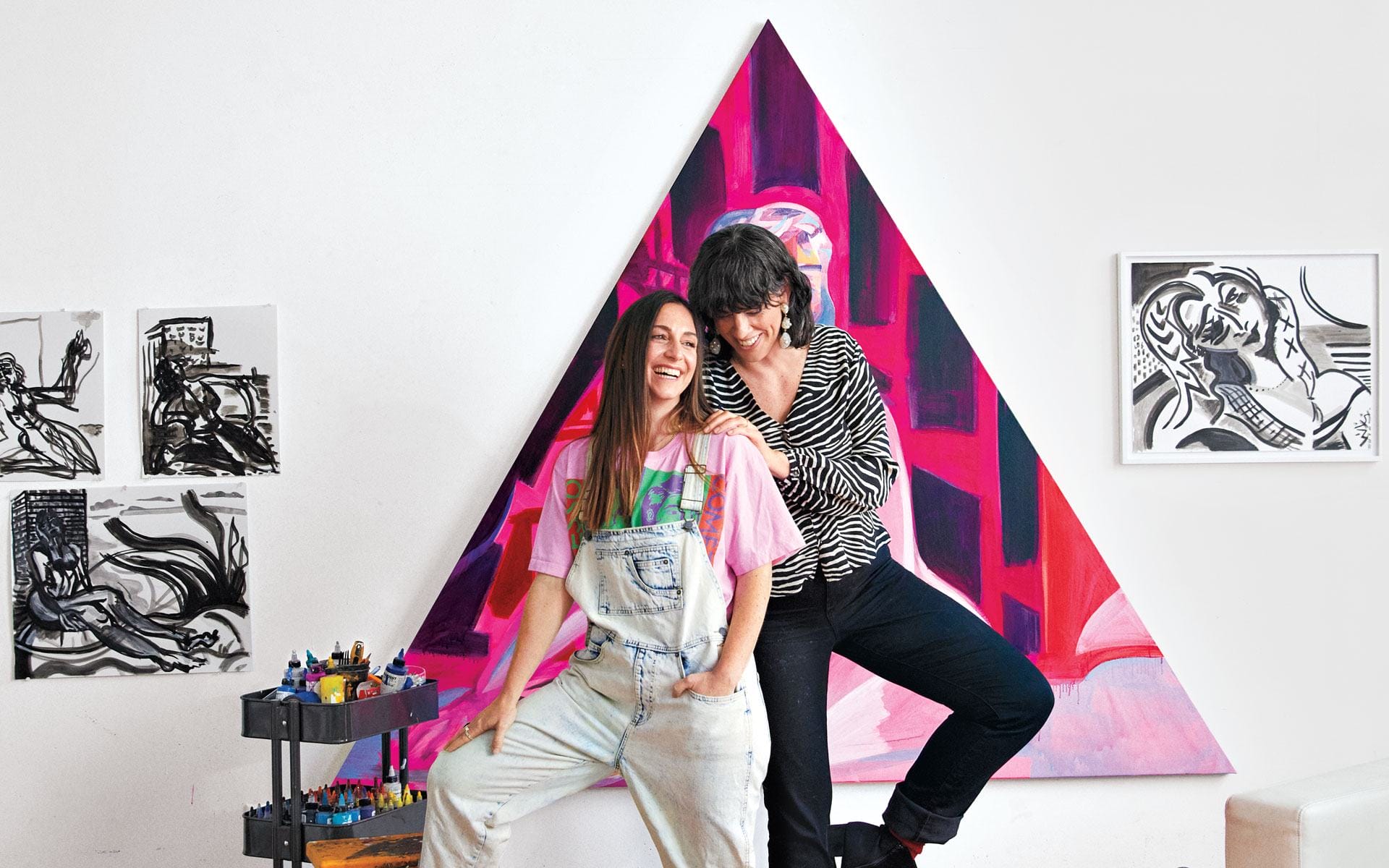 Two people smiling in an art studio with colorful triangular painting and artwork on the walls in the background.