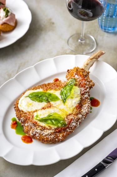 Breaded and fried dish with cheese and basil on a white plate, accompanied by a glass of red wine.