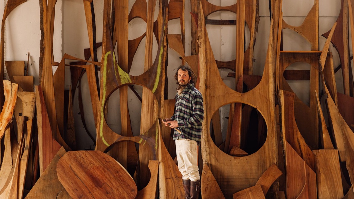 Man in a workshop surrounded by large wooden sculptures and abstract shapes, wearing a plaid shirt and boots.